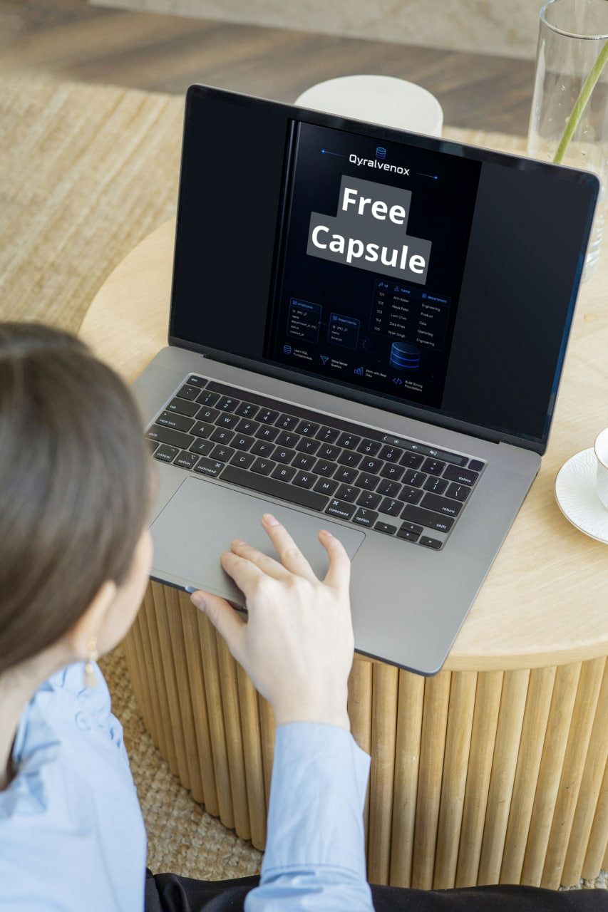 A person in a light blue shirt uses a silver laptop on a round wooden table in a modern indoor setting. The laptop screen displays the Dynavexos 'Free Capsule' interface.